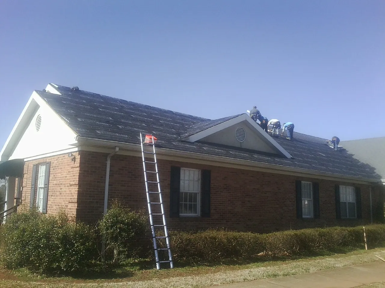 Crew tearing shingles off a brick ranch roof with a ladder set — Emergency Roof Repair work by our crew in Mc Clure