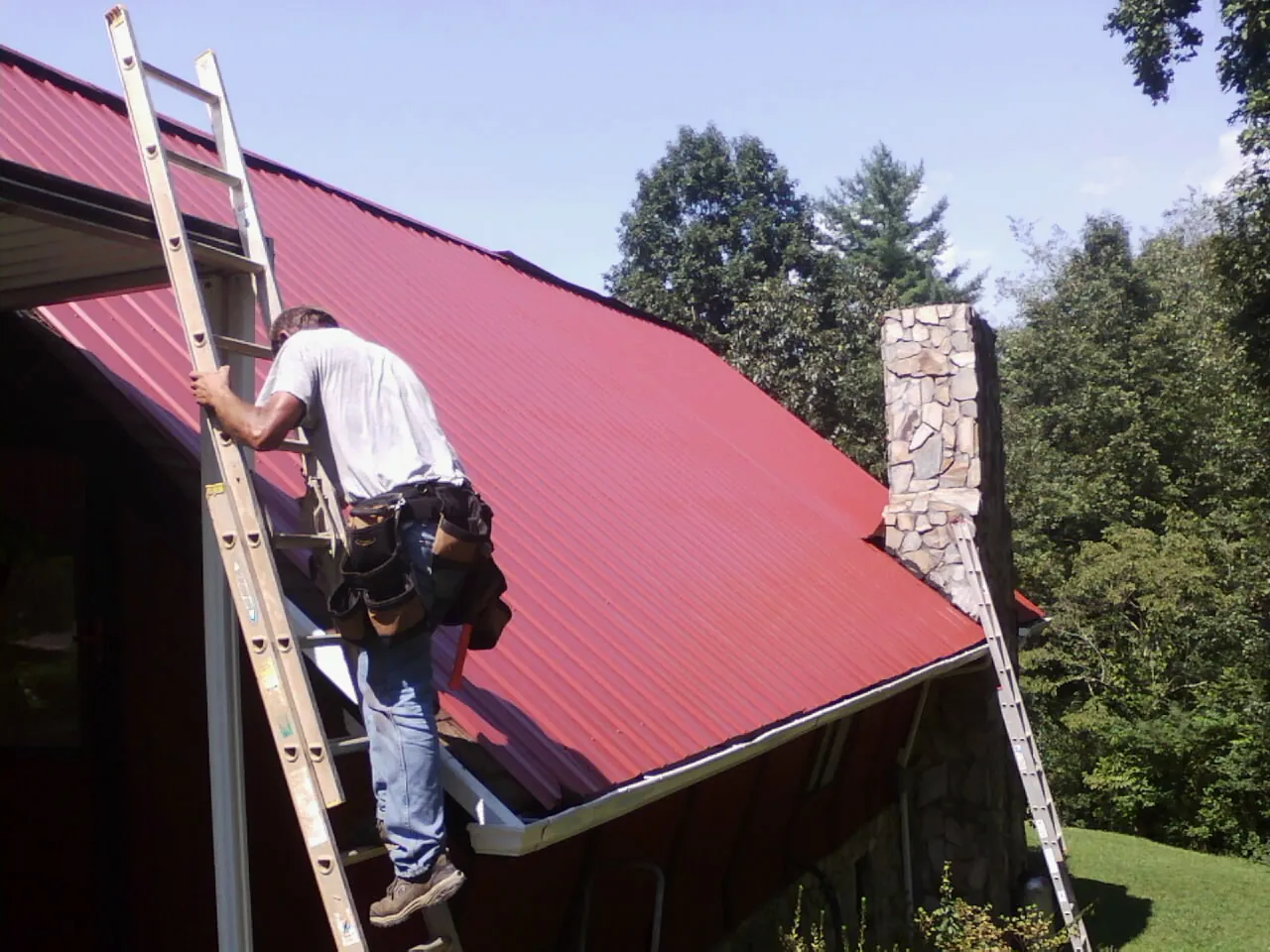 Roofer on a ladder beside a red metal roof and stone chimney — Soffit & Fascia Repair by Roofing Brothers in Mc Clure